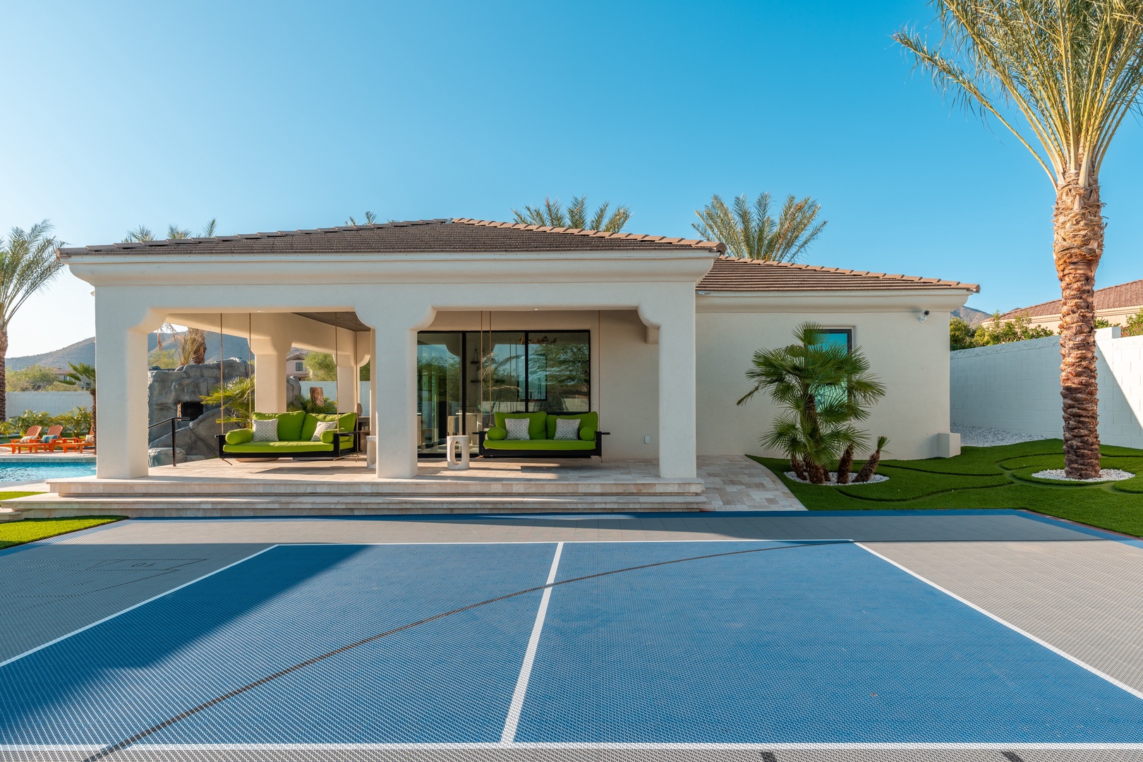 A modern desert-style villa features manicured lawns, palm trees, and a covered outdoor lounge. Taken during clear daylight, showing mountains in the distance.