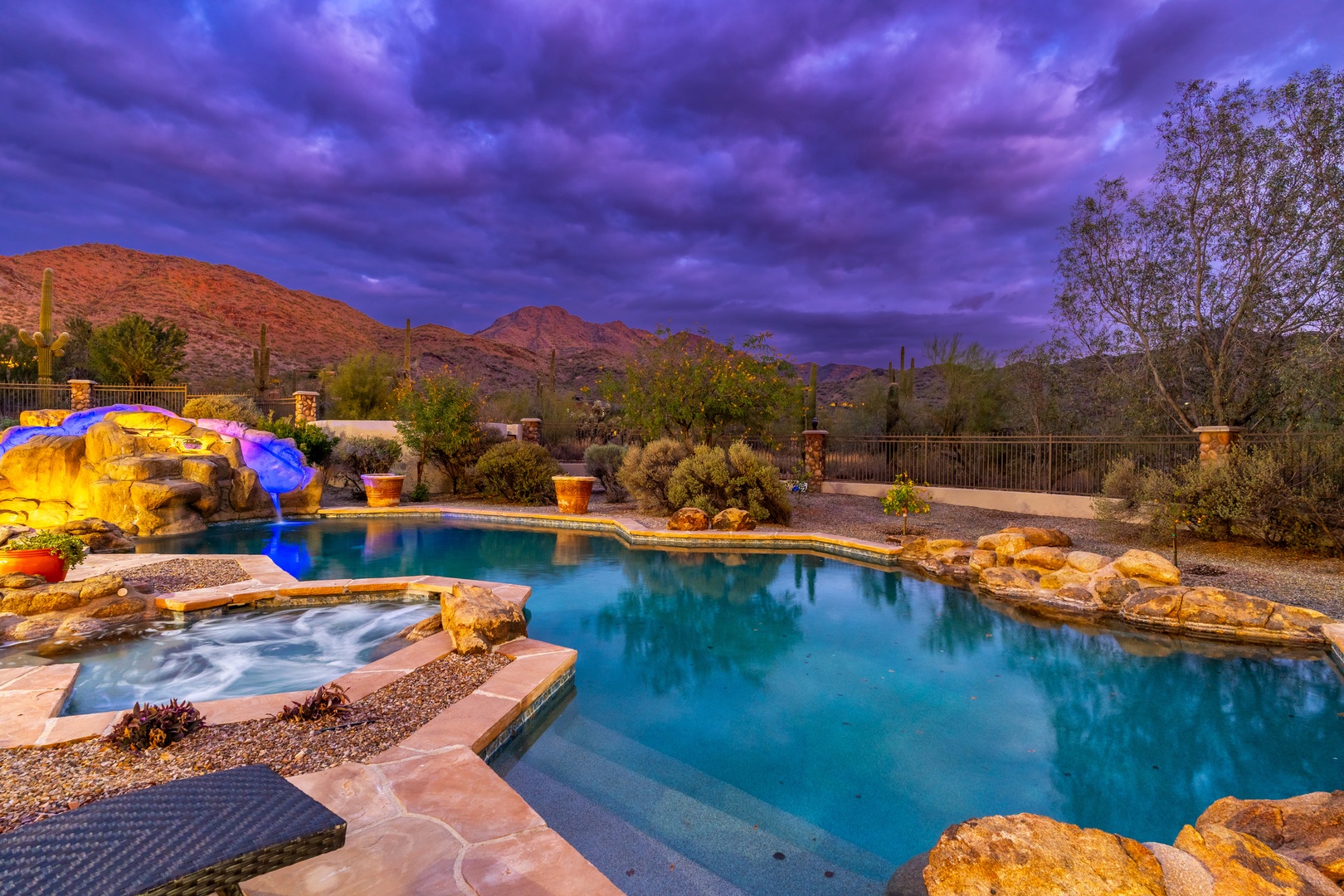 A stunning desert oasis awaits with crystal-clear pool and illuminated grotto, framed by majestic mountain views and dramatic twilight skies perfect for evening relaxation.