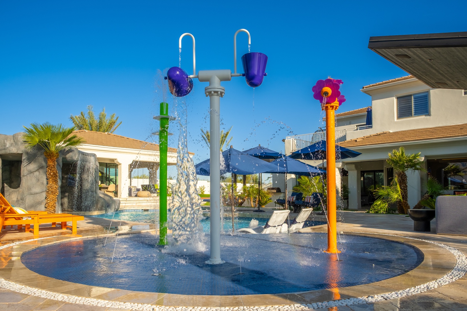 Colorful splash pad with interactive water features invites hours of fun and laughter under sunny blue skies, surrounded by tropical landscaping and pool loungers.