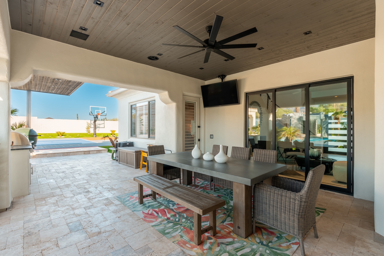 A covered outdoor dining area featuring a stylish table and seating, perfect for enjoying meals overlooking the pool and sunny backyard. Modern ceiling fan keeps you comfortable year-round.