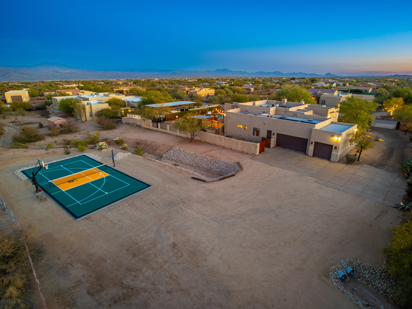 Aerial view of the property showcasing a private sport court, spacious outdoor grounds, and modern architecture nestled in a scenic desert neighborhood with mountain vistas at dusk.