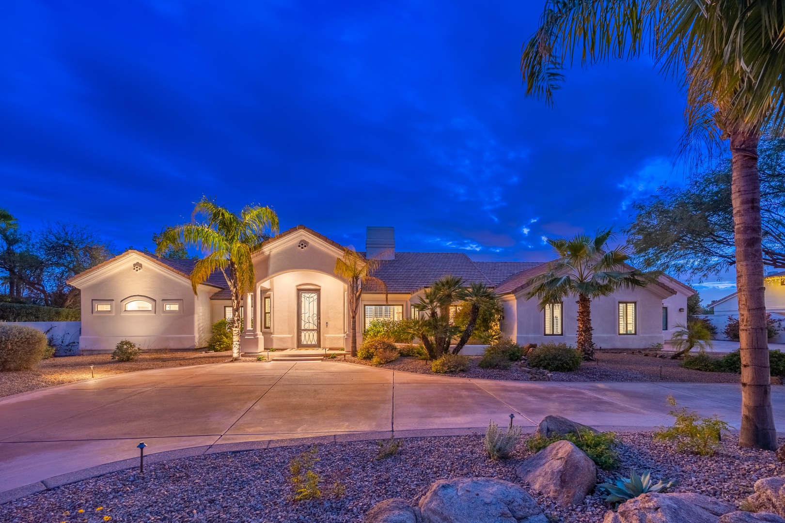 This elegant desert retreat greets you with warm lighting and stately palms as twilight settles in, offering a grand welcome to your tranquil vacation home.