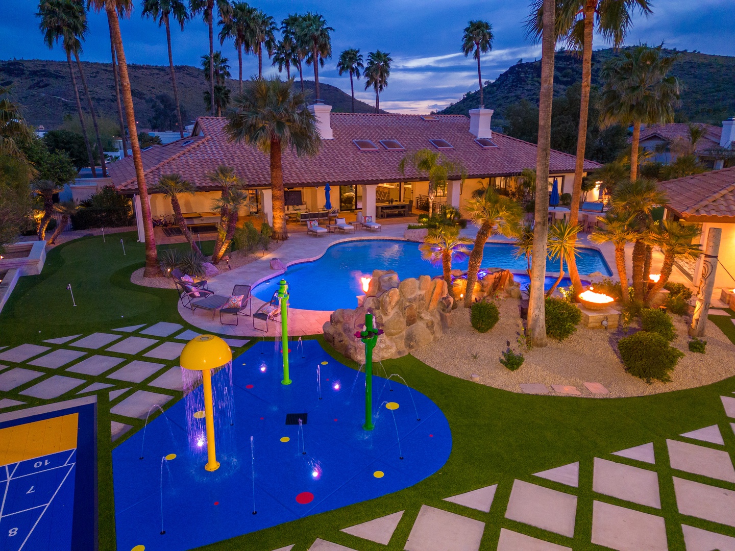 Aerial view at dusk of a sprawling estate with resort-style pool, integrated splash pad, manicured lawns, putting green, towering palms, and mountain backdrop—a private desert oasis for unforgettable gatherings.