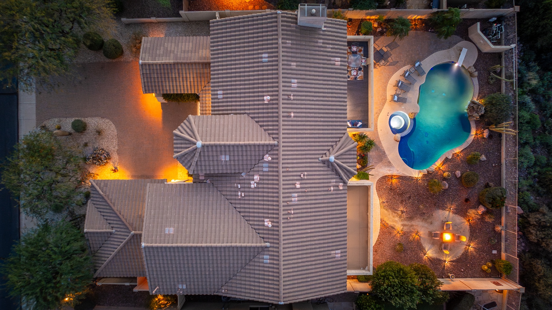 An aerial view captures the property at dusk, showcasing the sprawling tiled rooftops and a stunning free-form pool surrounded by ambient landscape lighting and lush natural greenery.
