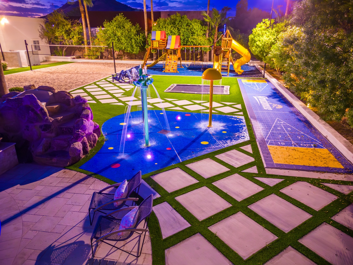 Your family will love this vibrant backyard resort featuring a splash pad, playground with yellow slide, hopscotch court, and plenty of space for outdoor fun under palm trees.