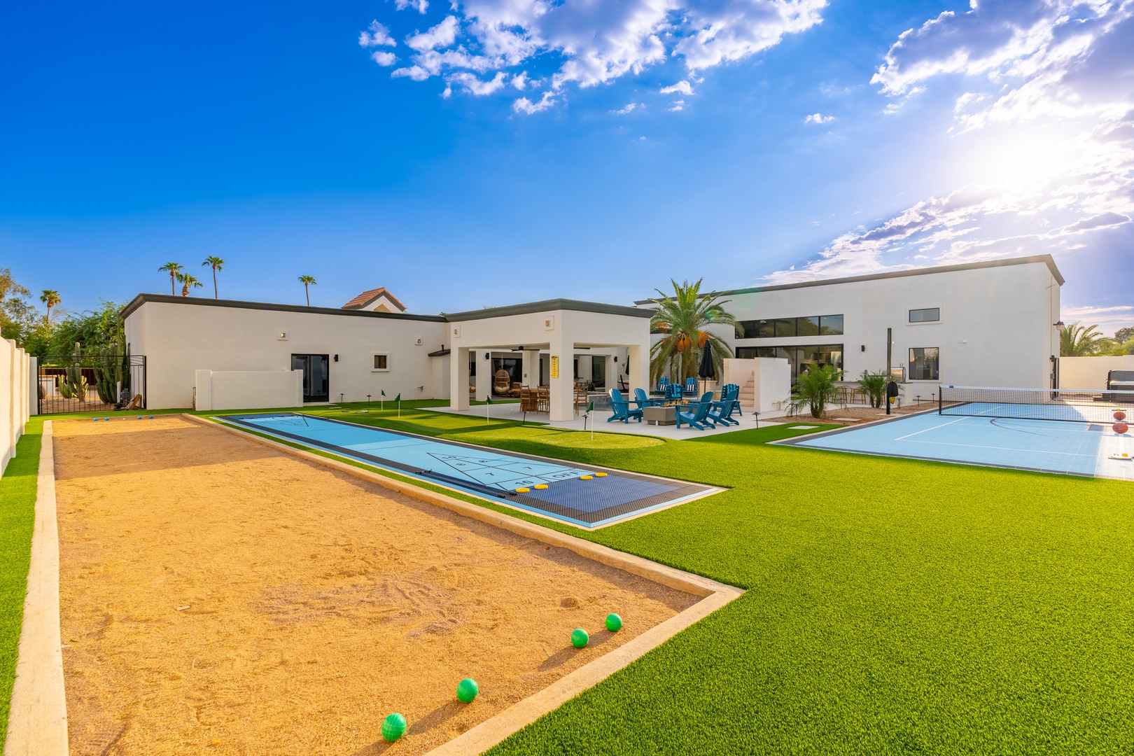 A contemporary vacation retreat featuring a sparkling pool, covered patio with blue lounge chairs, bocce court, and palm-studded lawn beneath wide Arizona skies—perfect for entertaining or unwinding.