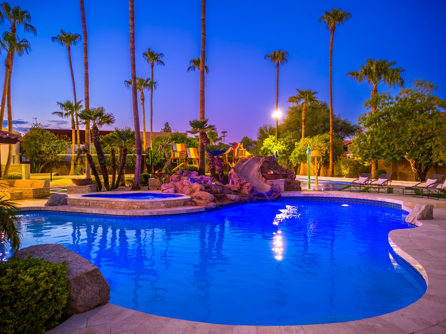 A stunning resort-style pool with a waterfall feature glows beneath twilight skies and swaying palms, creating a serene oasis for evening relaxation.