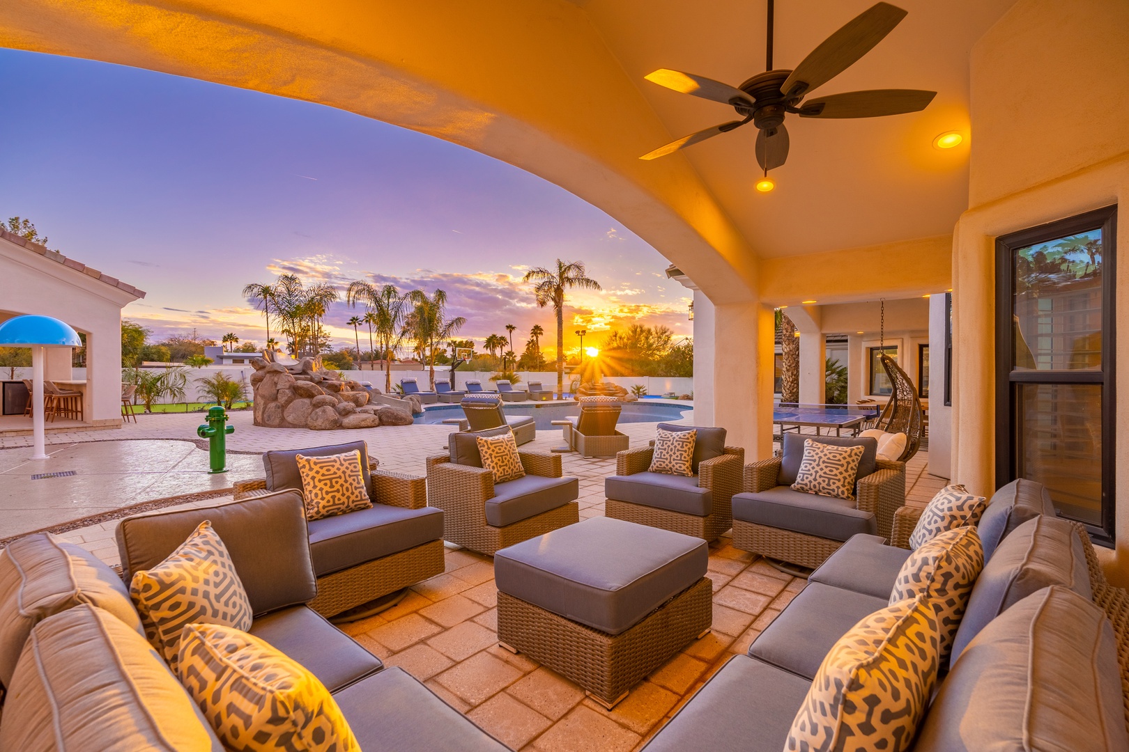 Sunset views frame this covered outdoor lounge, where you can relax on plush seating as golden light spills across the pool and swaying palms beyond.