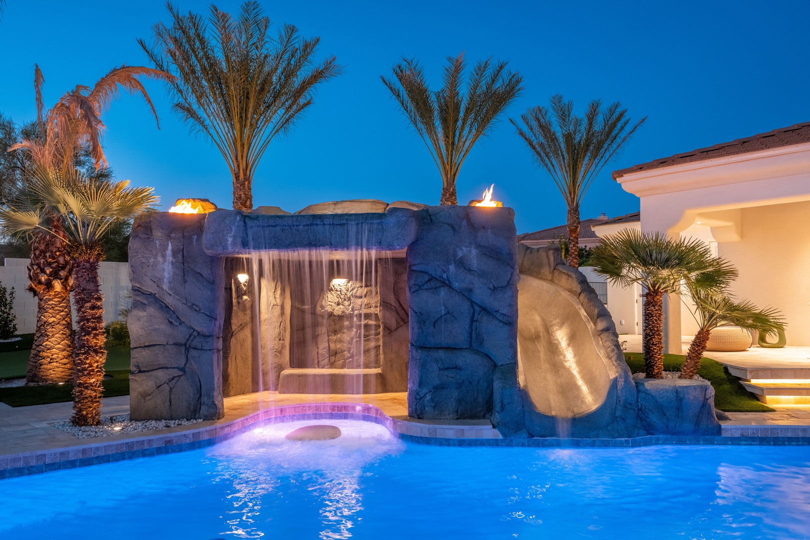 A luxurious pool at dusk features a stunning rock waterfall and built-in slide, surrounded by illuminated palm trees and tropical landscaping. The property's elegant exterior provides a perfect backdrop for evening relaxation.