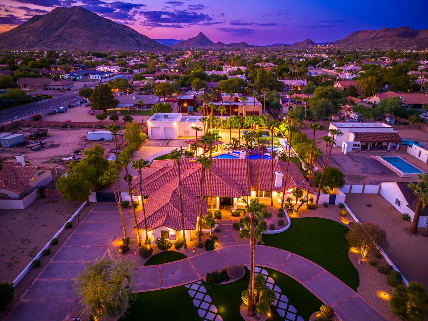 Aerial view captures the property nestled in a scenic desert neighborhood, framed by dramatic mountain peaks and a vibrant twilight sky transitioning from purple to orange.