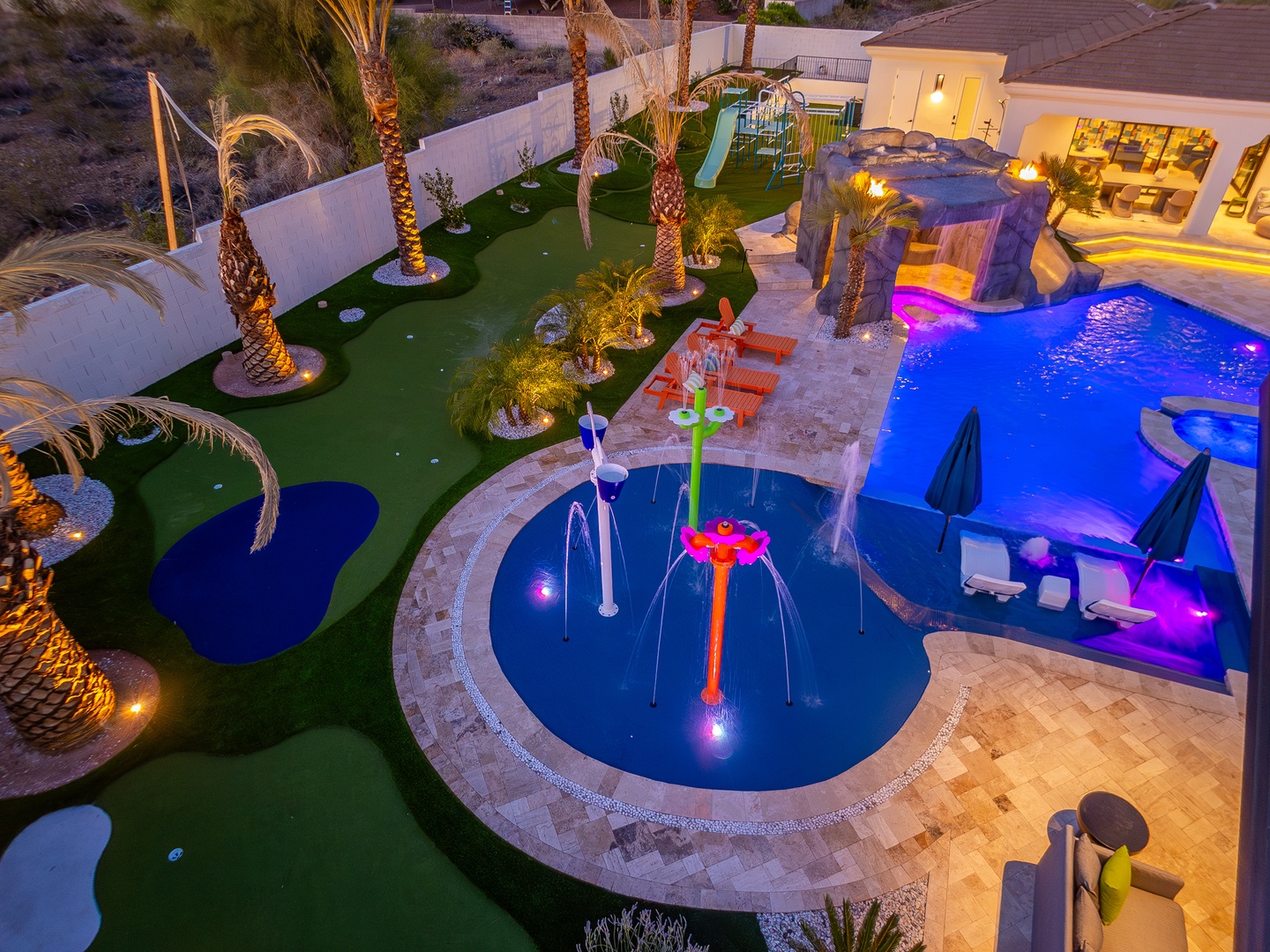 Aerial view of a resort-style backyard featuring a mini-golf course, swimming pools with waterfalls and colorful lighting, splash pad, and tropical palm landscaping at dusk.