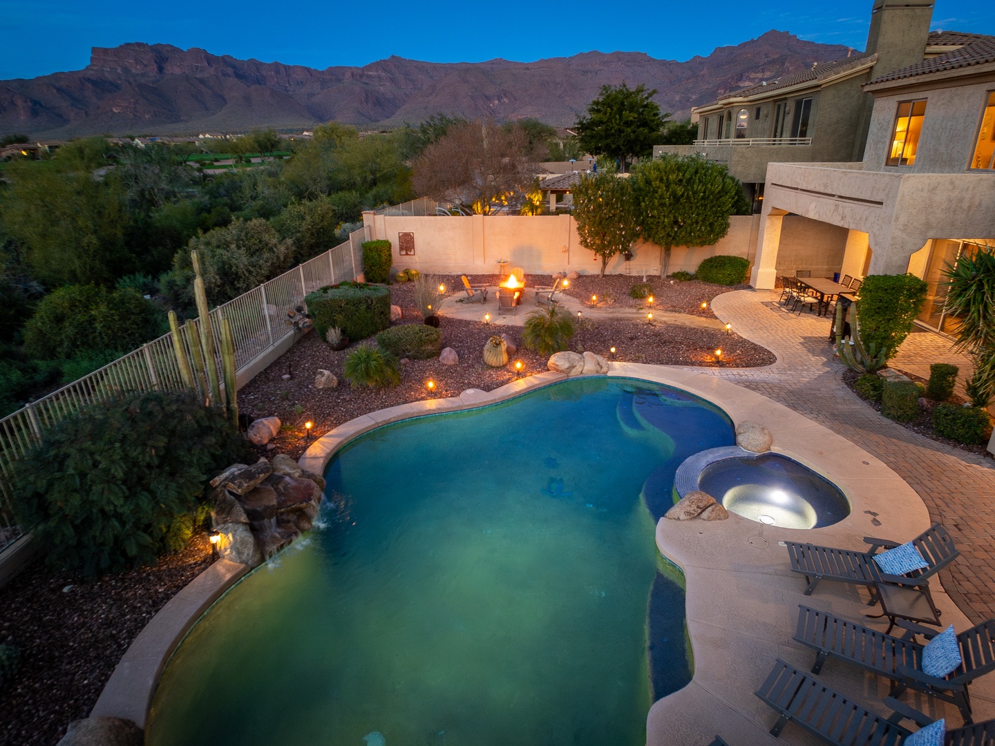 Aerial view captures the property's expansive backyard oasis at twilight, featuring a glowing pool, spa, firepit seating areas, and desert landscaping framed by mountain views.