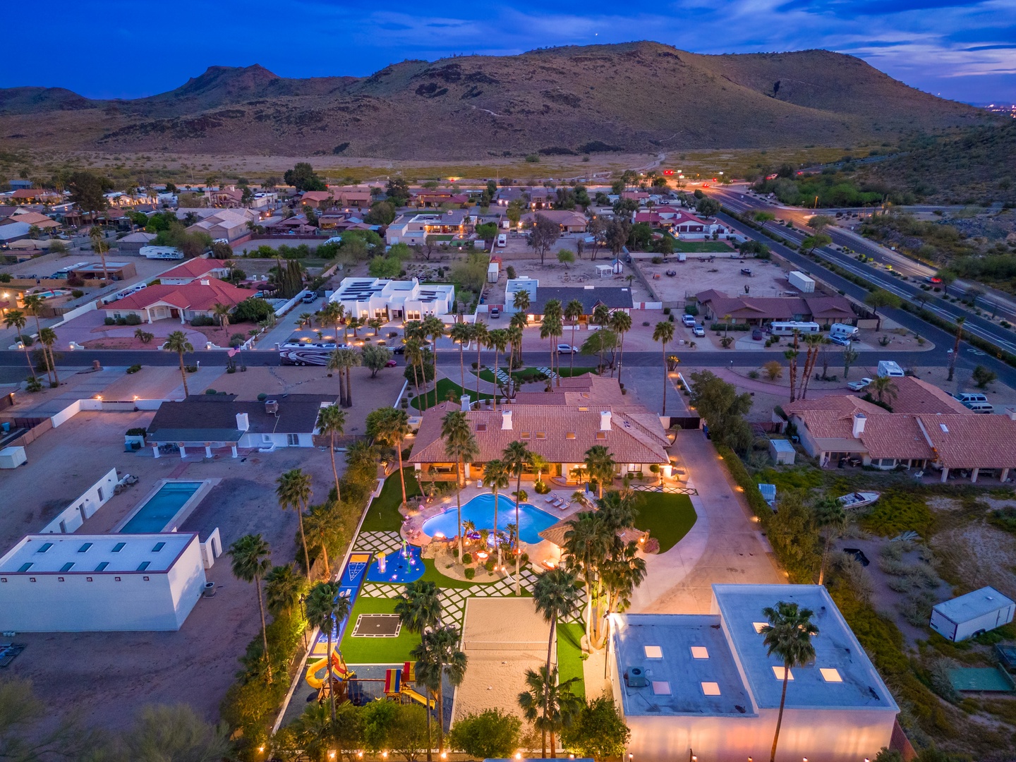 An aerial view captures the desert-oasis property nestled in a mountain-framed neighborhood, featuring its illuminated pool and landscaped grounds beneath twilight skies.