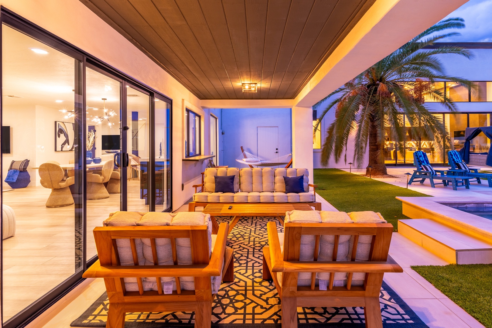 Modern covered patio at dusk featuring stylish lounge seating, warm ambient lighting, poolside views, and tropical palm trees just beyond your outdoor retreat.
