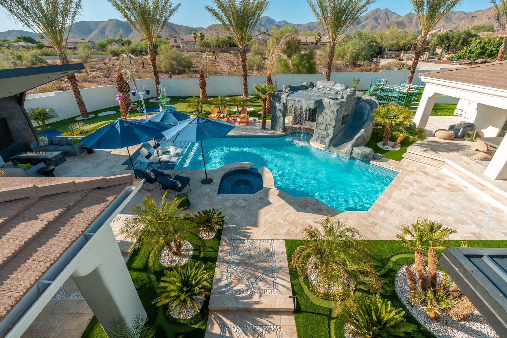 This stunning desert oasis features a resort-style pool with a waterslide rock formation, surrounded by lush landscaping and mountain views, with multiple lounge areas for relaxation.