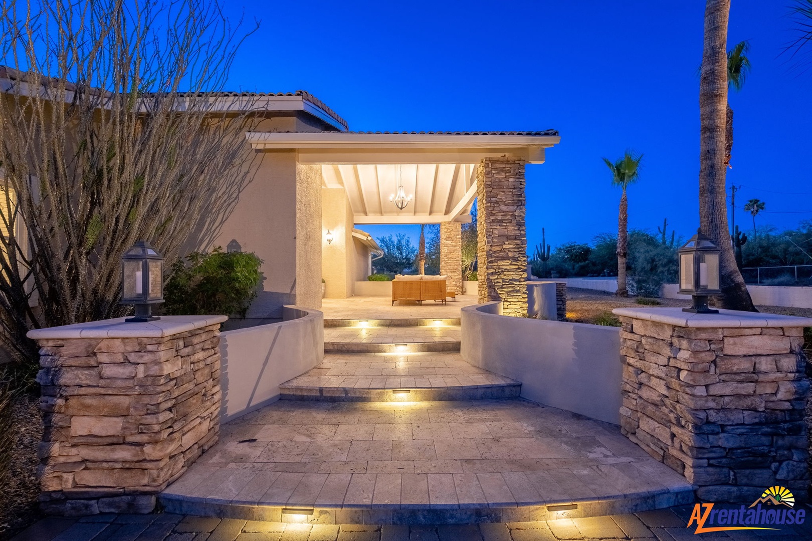 The property's welcoming entrance features elegant stone columns, ambient lighting, and desert landscaping, creating a warm arrival experience at dusk beneath a vibrant blue sky.