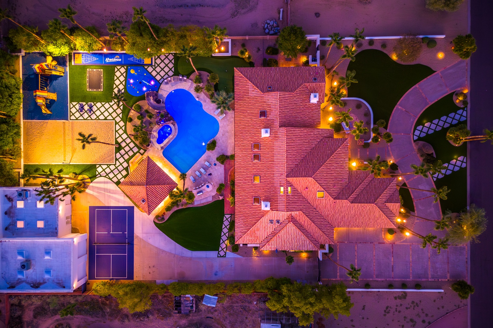 Aerial view of the resort property at night, showcasing the main building with terracotta roofing, illuminated swimming pool, tennis court, lush tropical landscaping, and surrounding recreational facilities.