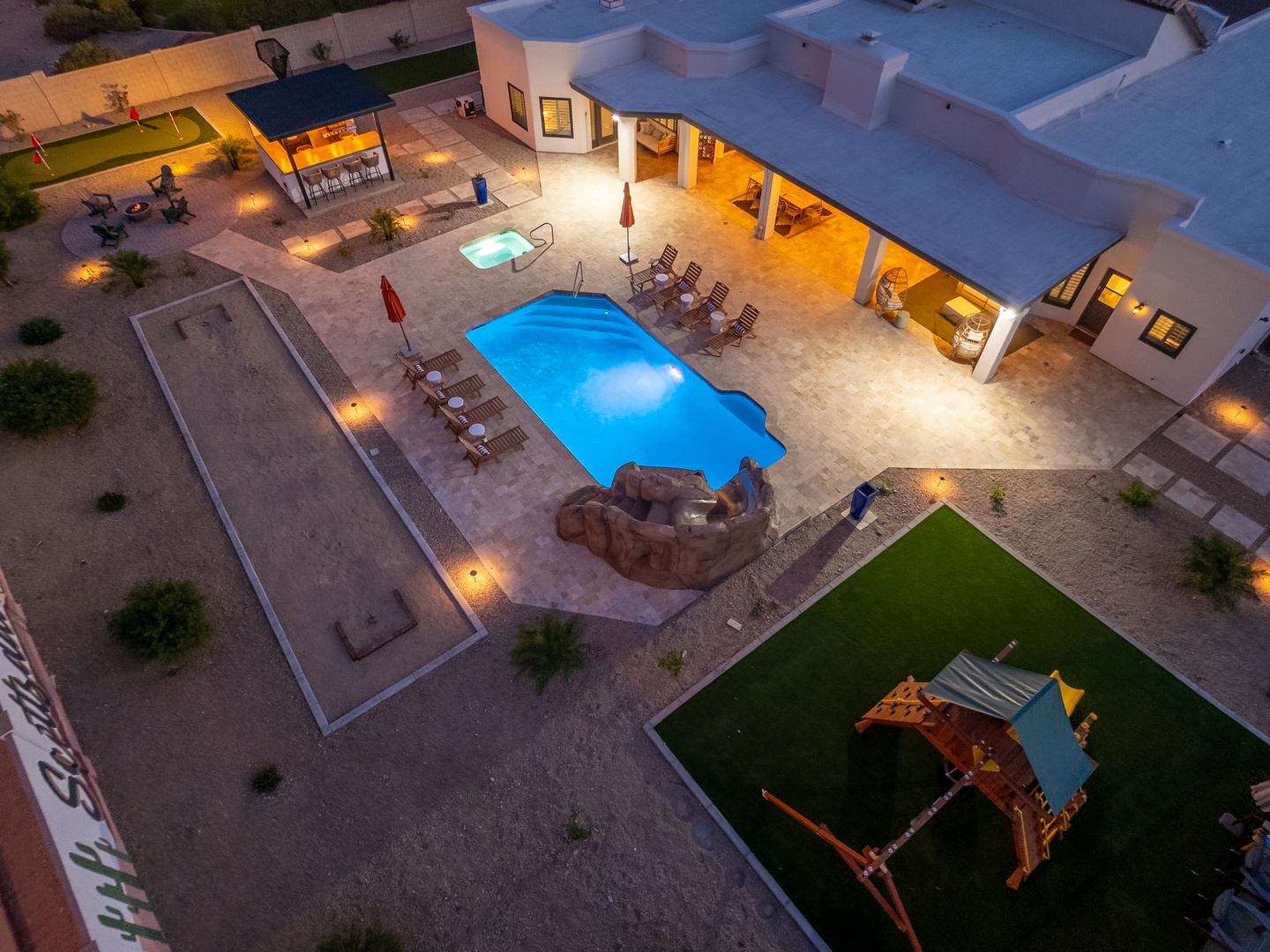 Aerial view of the spacious property at twilight, featuring a glowing pool, children's play area, sports court, and ample outdoor lounging spaces surrounded by desert landscaping.