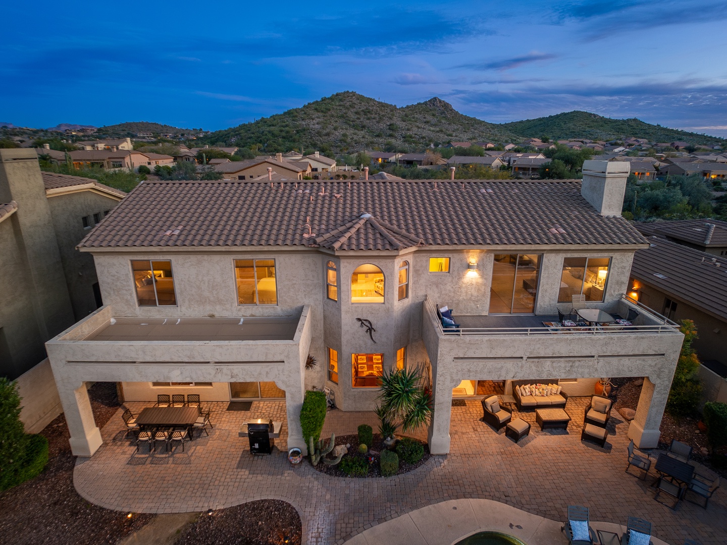 Aerial view of a spacious desert property featuring illuminated interiors, covered outdoor living areas, and mountain surroundings at dusk.