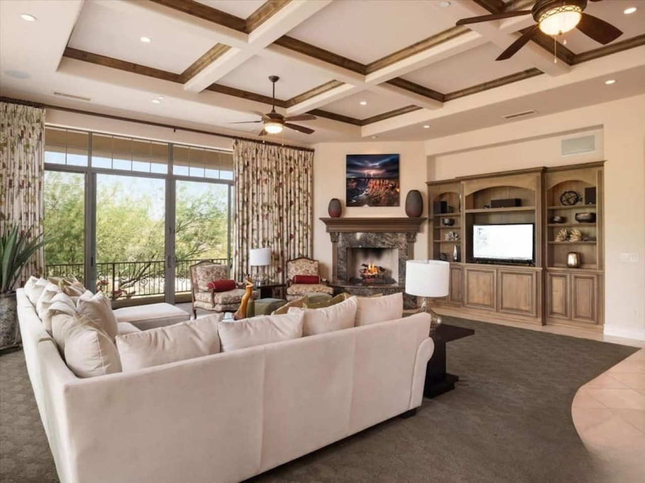 Relax in this elegant living room with soaring coffered ceilings, a cozy fireplace, and plush seating overlooking lush greenery through expansive windows—your perfect retreat awaits.