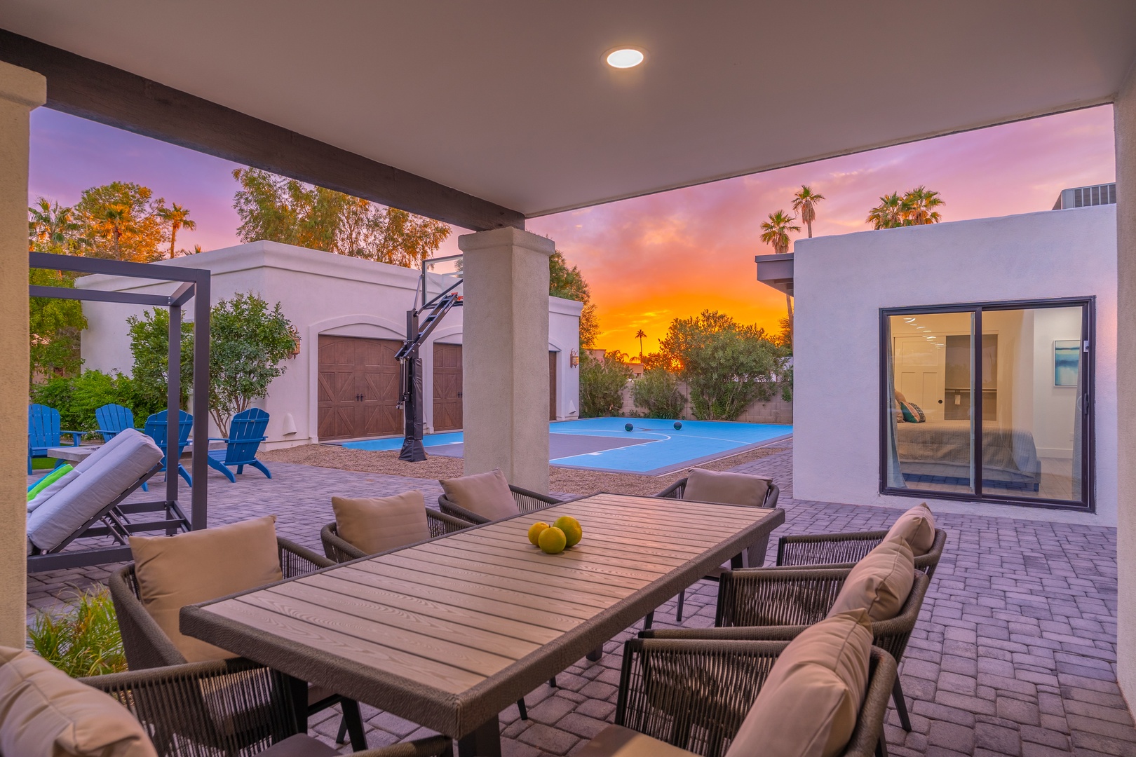 A stunning desert sunset paints the sky behind this outdoor entertainment space, where covered dining for eight awaits alongside vibrant blue Adirondack chairs and a sparkling pool beyond.