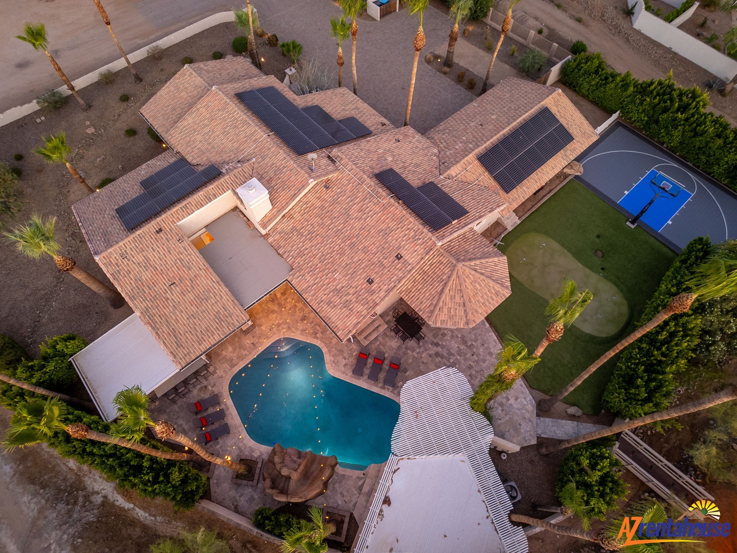 Aerial view of a spacious desert retreat featuring a private pool, outdoor sports court, solar panels, and palm-lined landscaping in a quiet residential neighborhood.