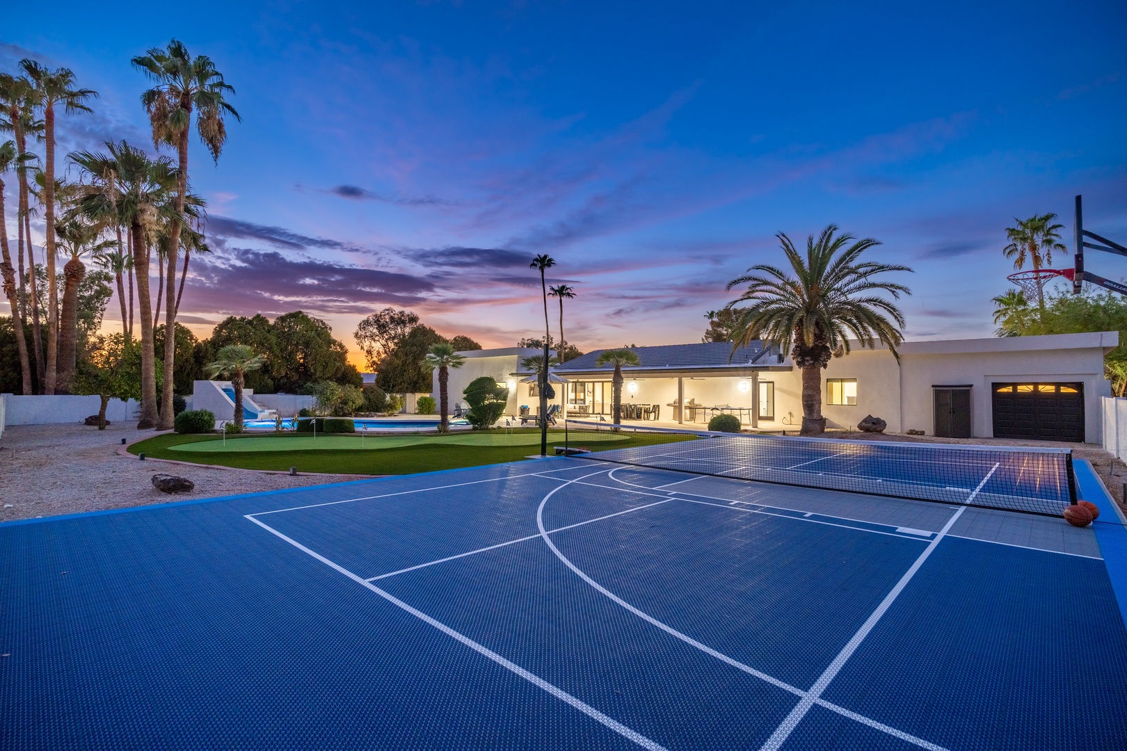 A modern desert oasis photographed at twilight, with a private basketball court surrounded by manicured grounds, palm trees, and a sleek main residence beneath a vibrant pink and blue sky.