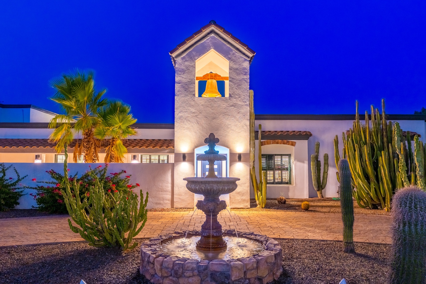 A striking Spanish-style bell tower rises above a courtyard fountain, surrounded by desert palms and towering cacti, illuminated against the twilight sky.
