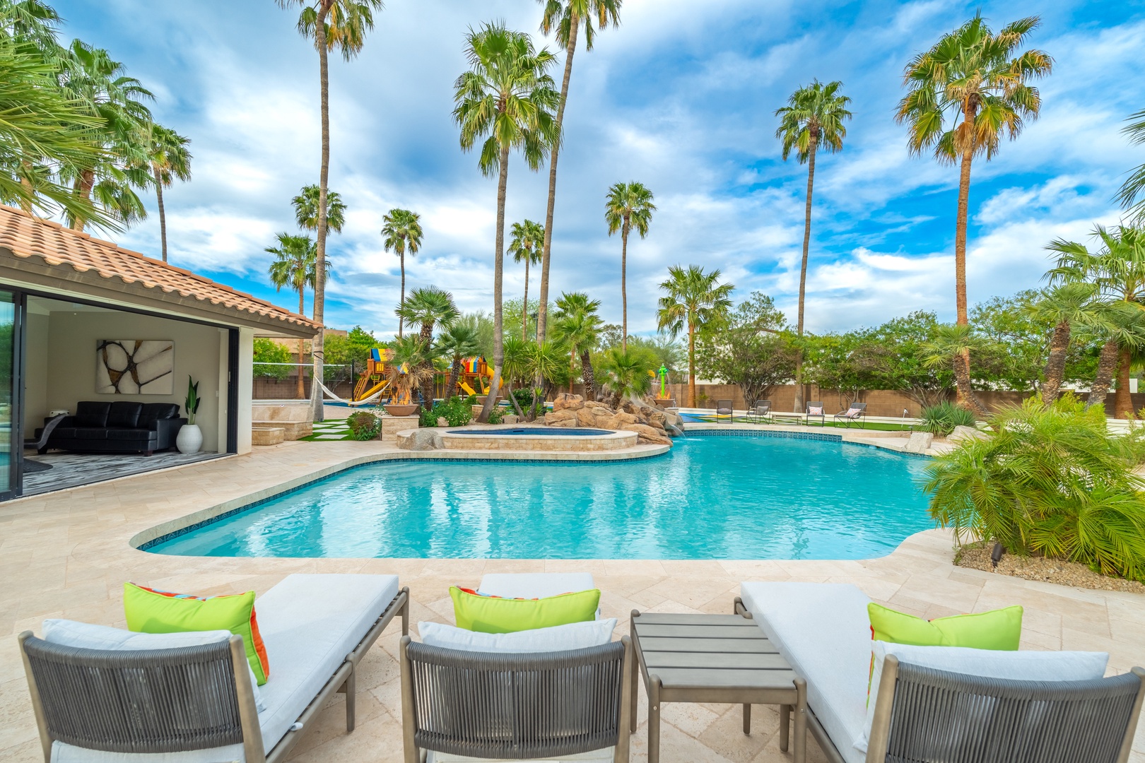 The sparkling pool features a unique curved design and integrated hot tub, surrounded by towering palms and inviting loungers—perfect for your desert oasis getaway.