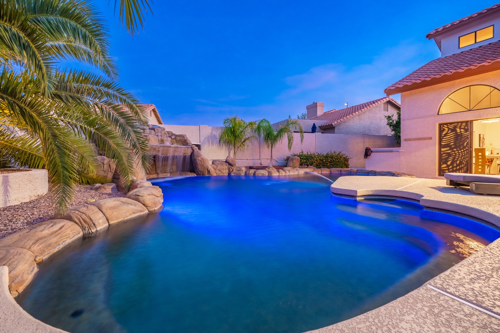 A twilight oasis awaits: glowing pool waters, tropical palms, and a cascading waterfall create your private resort-style escape under the desert sky.