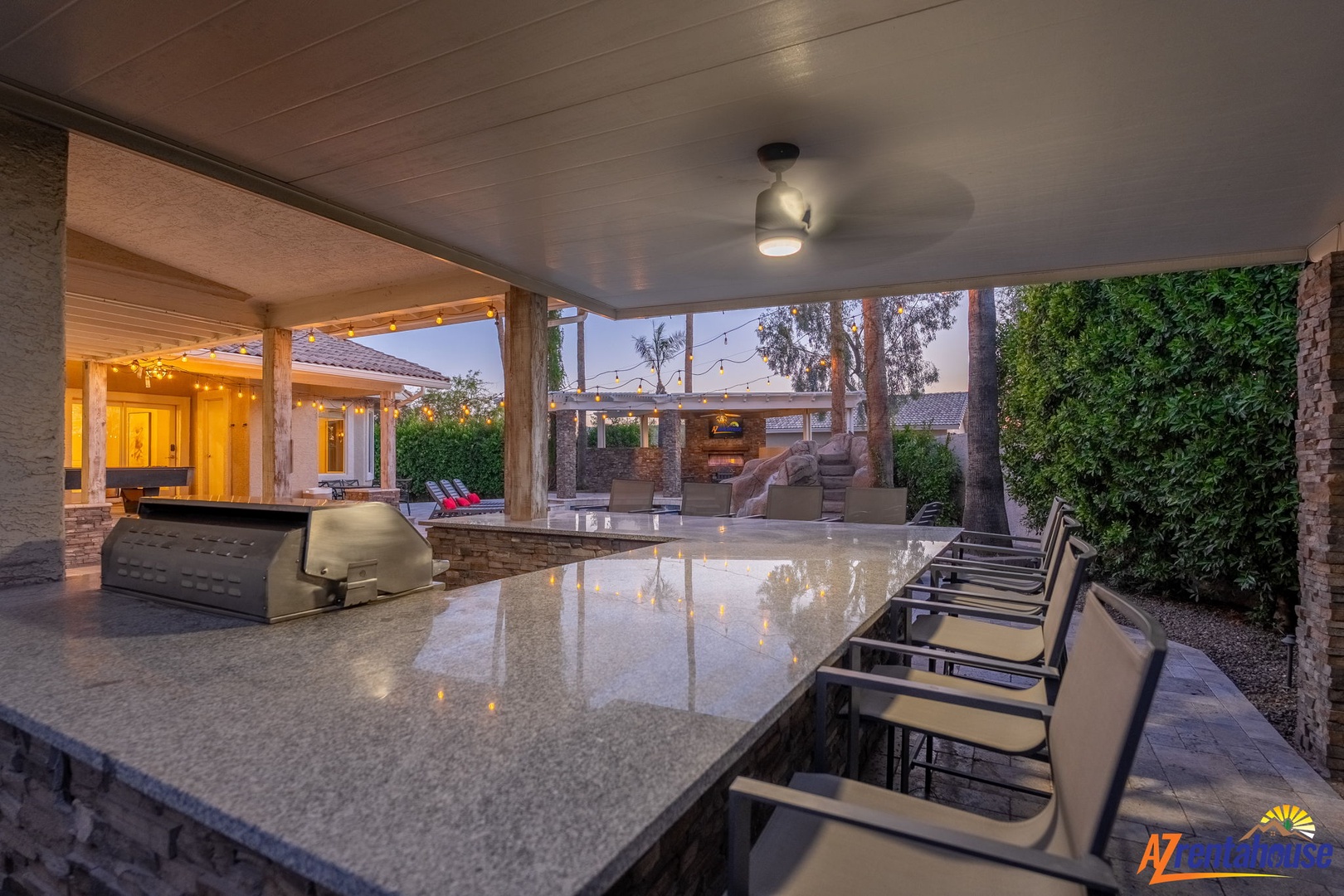 An expansive outdoor kitchen and bar area with granite countertops, built-in grill, and multiple seating stations opens to a covered patio perfect for evening entertaining under string lights.