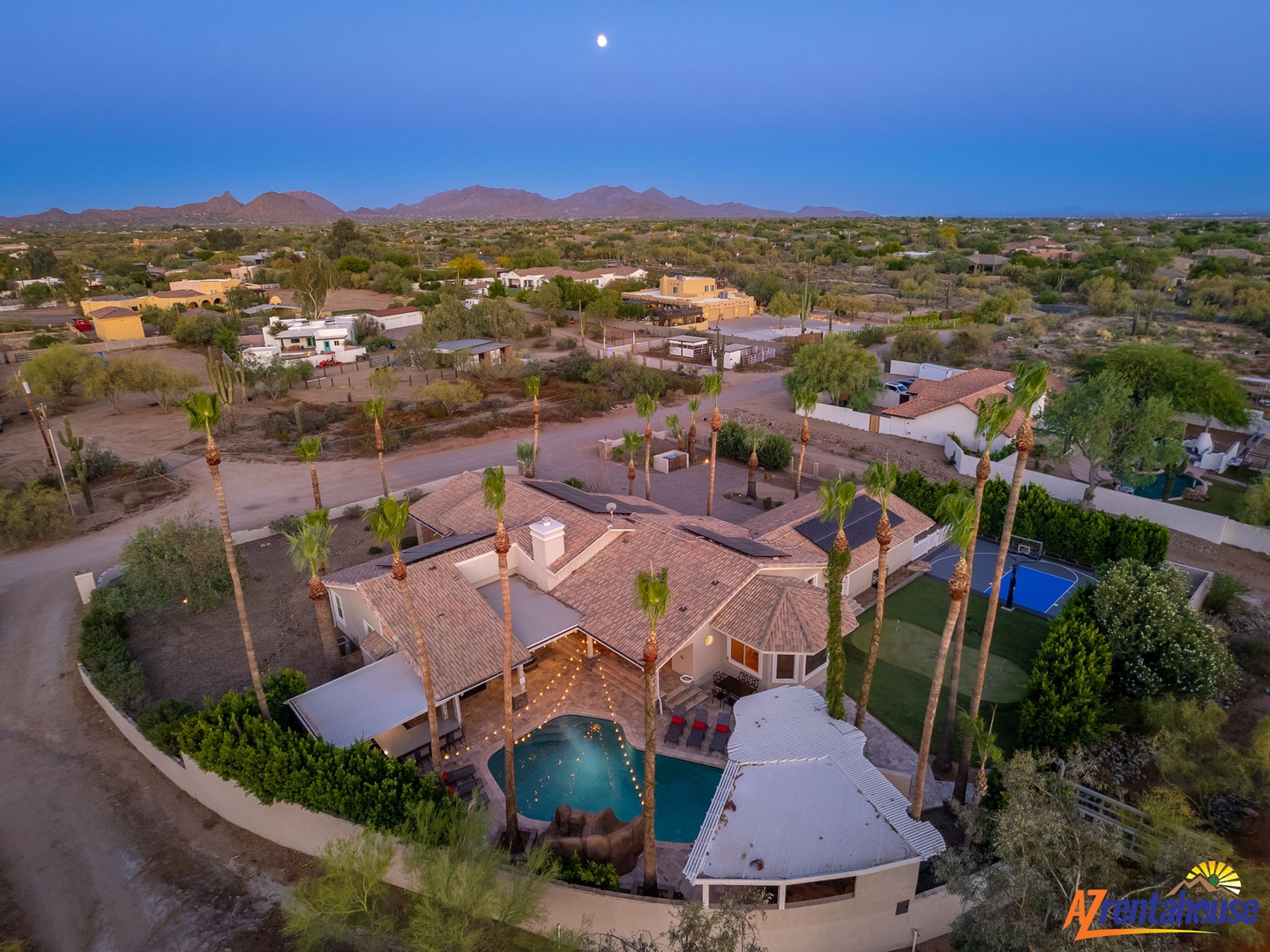 Aerial view of a desert retreat with private pool surrounded by towering palms, set against rugged mountain ridges and evening sky.
