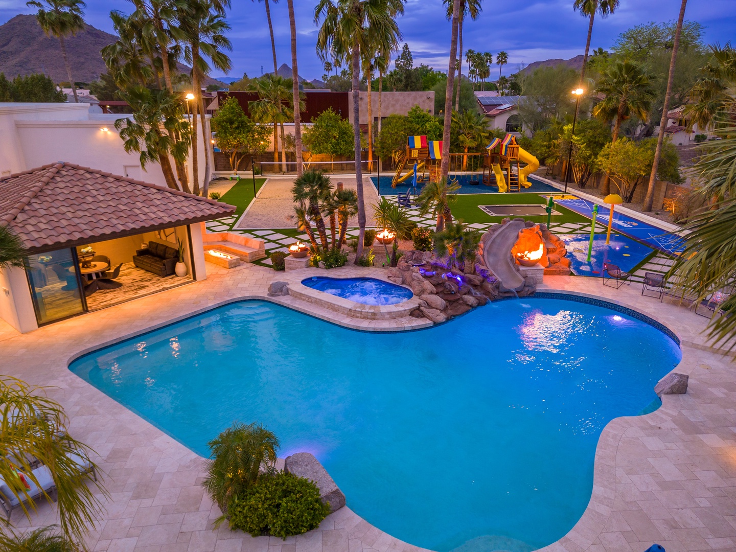 A resort-style backyard oasis featuring a sparkling pool with an integrated hot tub, illuminated waterslides, and mountain views at dusk.
