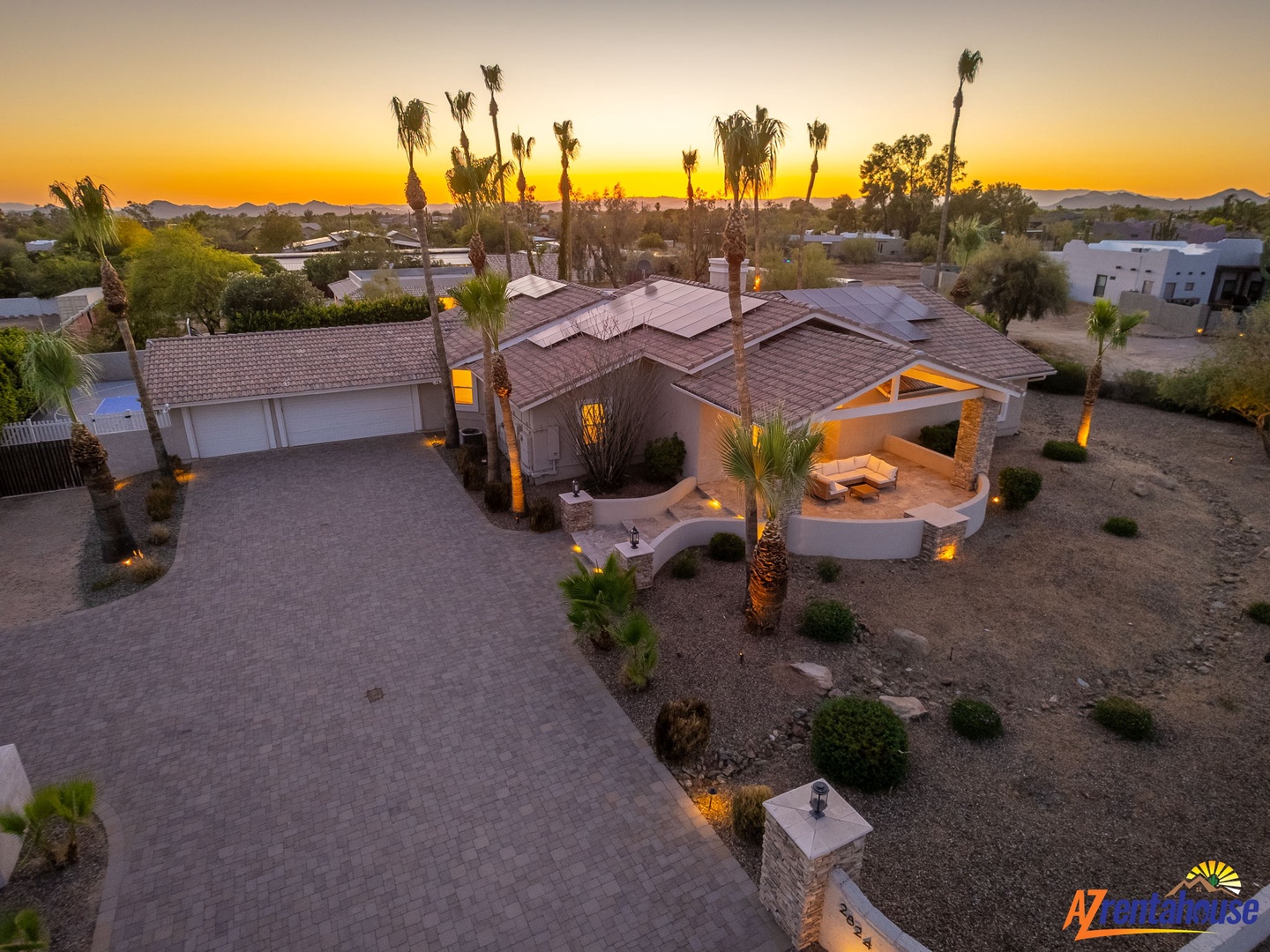 Aerial view captures the property at dusk, with warm lighting highlighting the desert landscaping and palm trees. Golden sunset illuminates the surrounding neighborhood and distant mountain ranges.