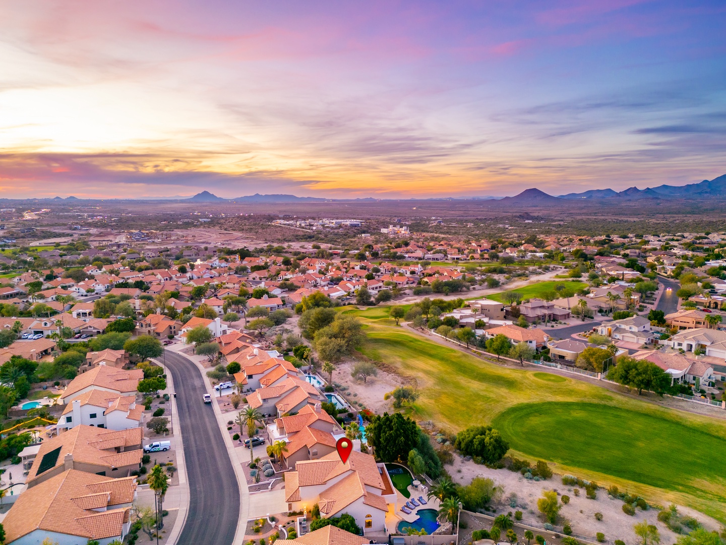 An aerial view captures the property's location within a residential neighborhood, bordered by a lush golf course and surrounded by stunning desert mountain vistas at sunset.