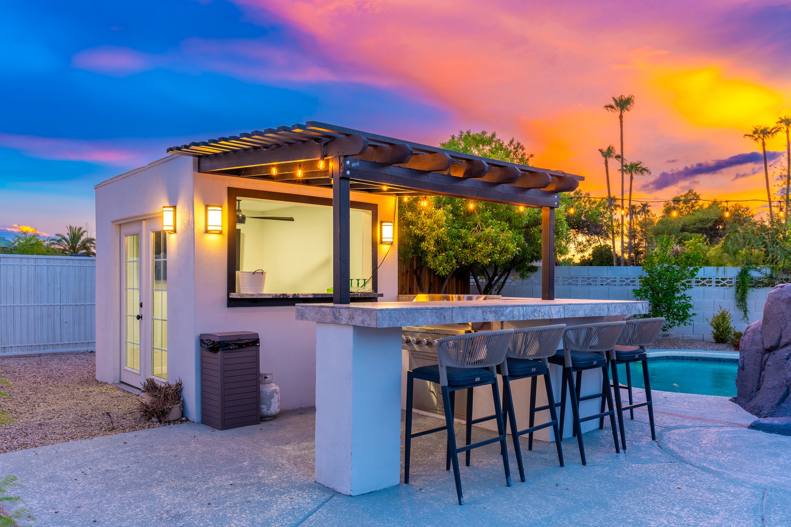 A stunning outdoor bar and dining area glows warmly beneath a dramatic desert sunset, with lush palms framing the space and your private pool just steps away.