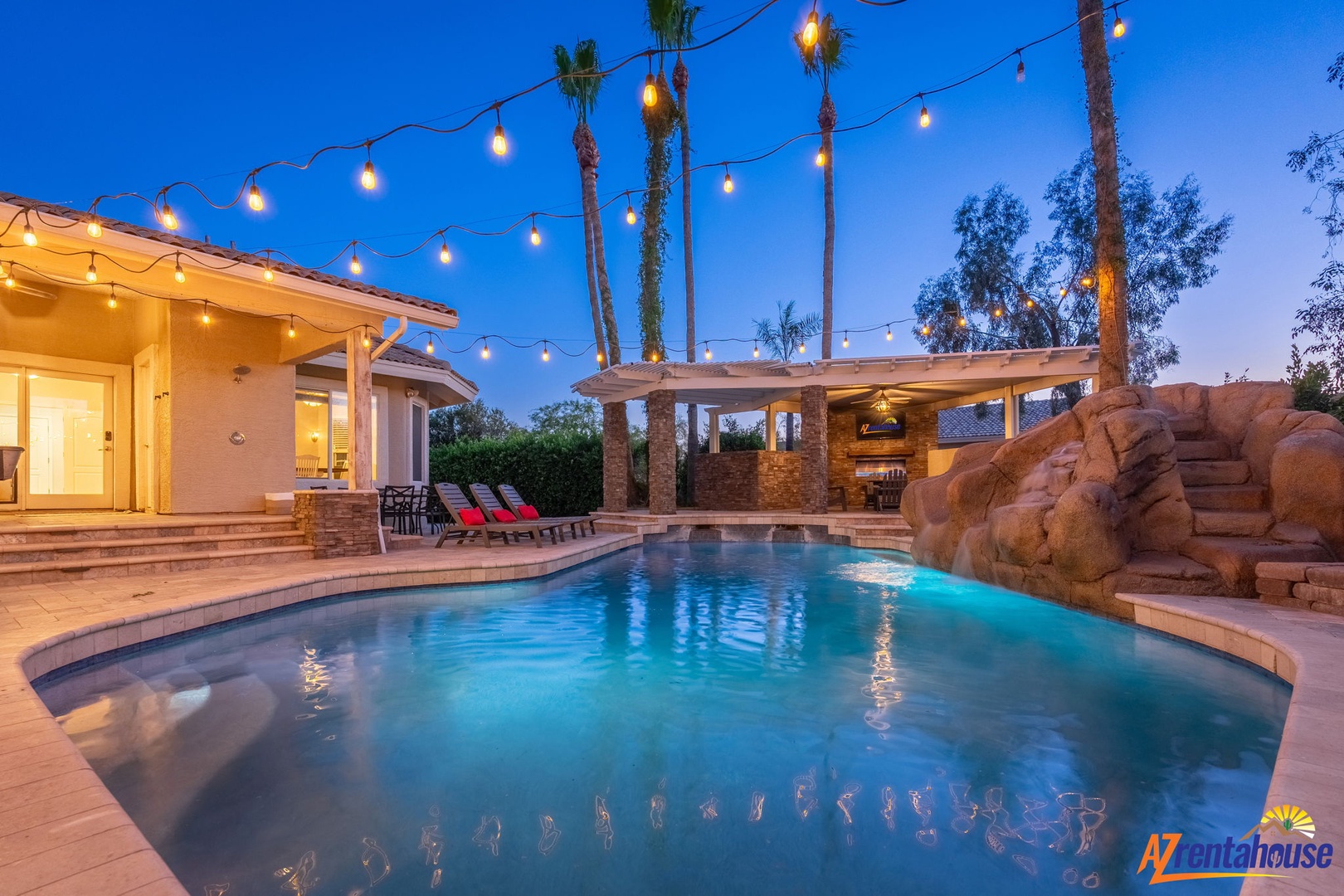This desert resort-style backyard offers an illuminated pool with a cascading rock waterfall, shaded lounges, and string-lit palms creating a magical twilight setting for your evening relaxation.