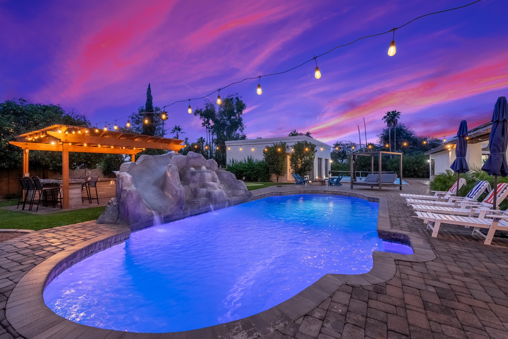 An illuminated pool area with waterfall feature, string lights, and covered patio under a dramatic sunset sky creates a serene evening atmosphere in the surrounding neighborhood.