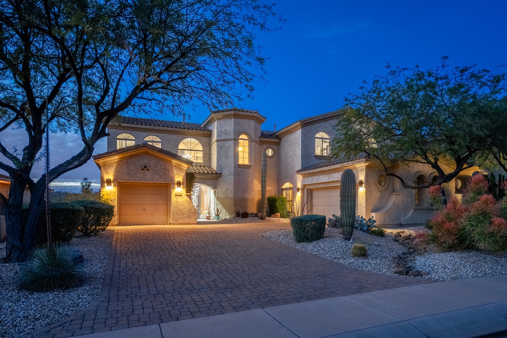 Stunning Spanish-style villa nestled among mature desert trees, featuring classic stucco arches, warm evening lighting, and a welcoming brick driveway surrounded by native Sonoran landscaping.