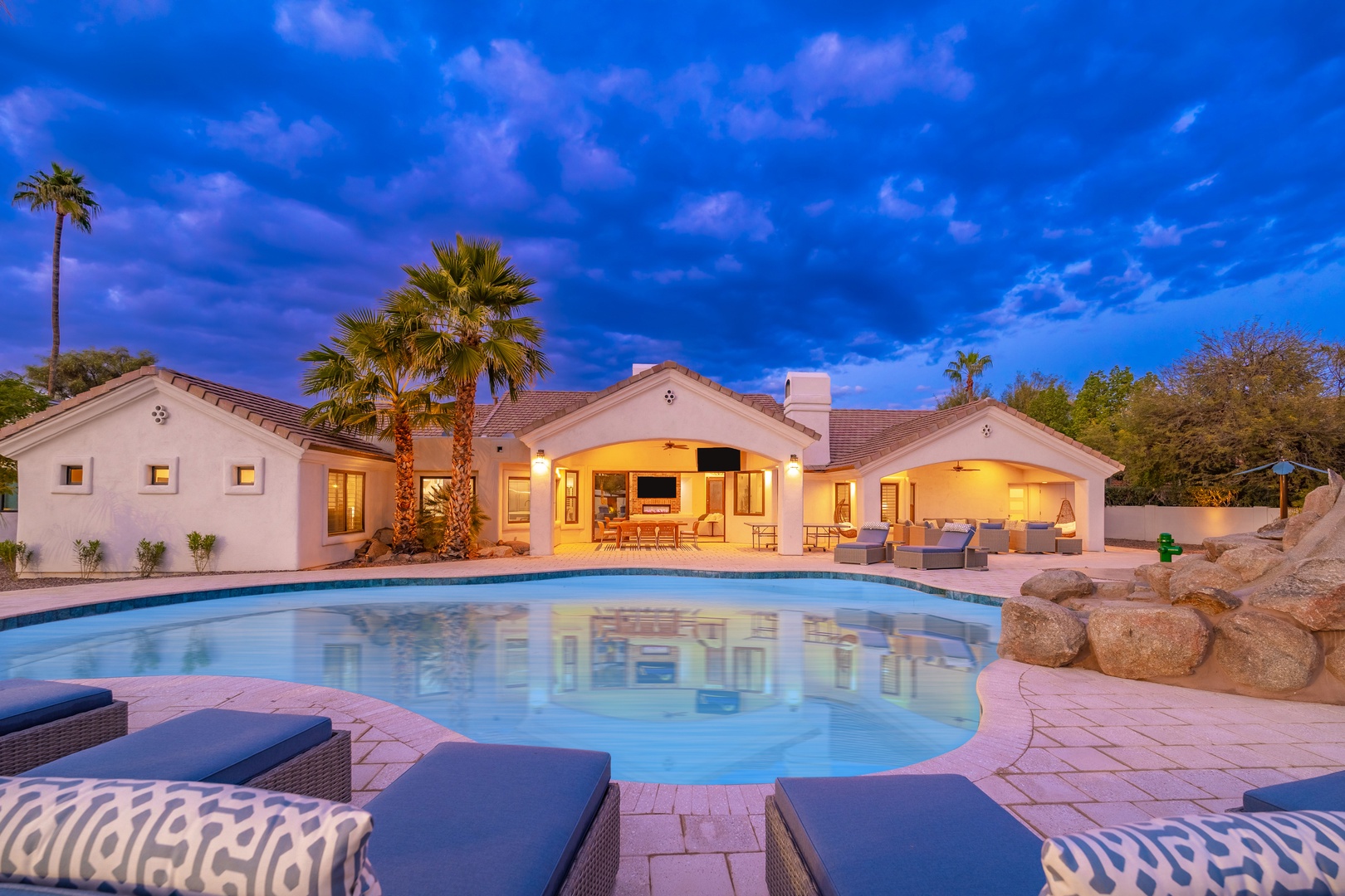 Elegant desert retreat featuring a sparkling pool surrounded by palm trees, spacious covered patio with comfortable seating, and breathtaking twilight skies creating a serene outdoor oasis.