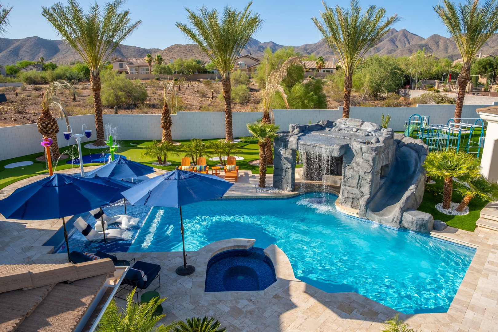 A stunning private oasis boasts a crystal-blue pool with waterfall grotto, spa, shaded lounging areas, lush palm trees, and dramatic mountain views—perfect for your desert escape.