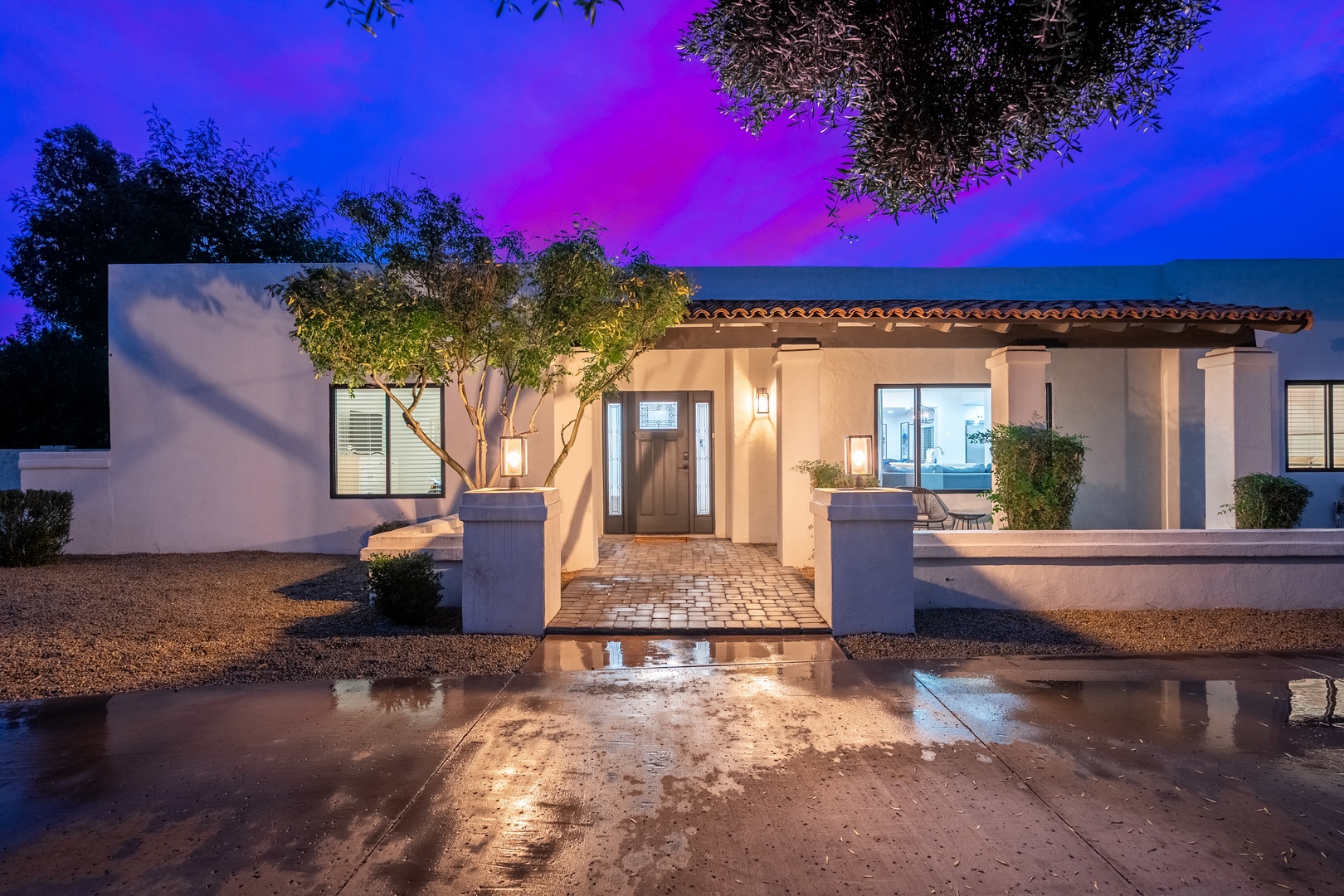 Elegant desert villa entrance illuminated at twilight, featuring terra-cotta tile roofing, mature landscaping, and dramatic purple skies creating a stunning arrival experience.