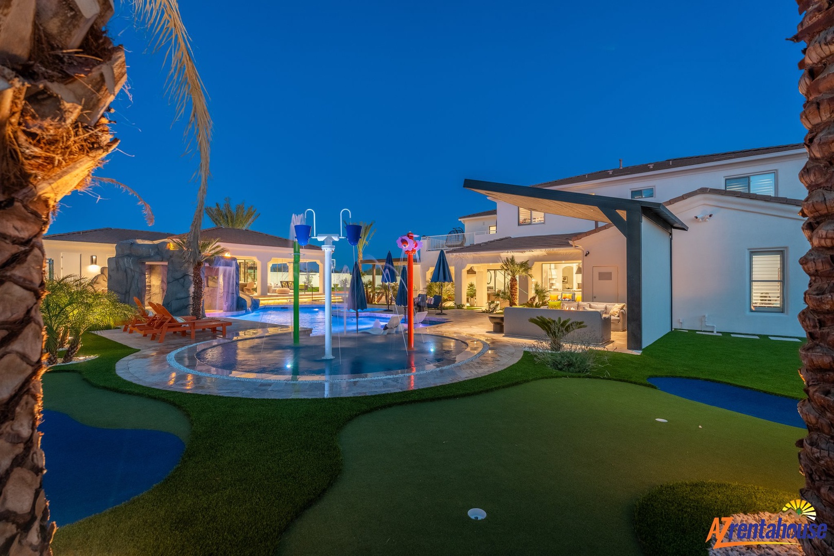 Modern villa with illuminated pool featuring colorful water features and mini golf course under palm trees at dusk.