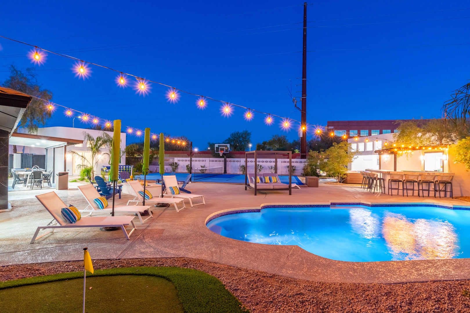 A stunning twilight scene showcases a resort-style backyard with a sparkling pool, loungers with striped cushions, putting green, basketball hoop, covered patio, and festive string lights overhead.