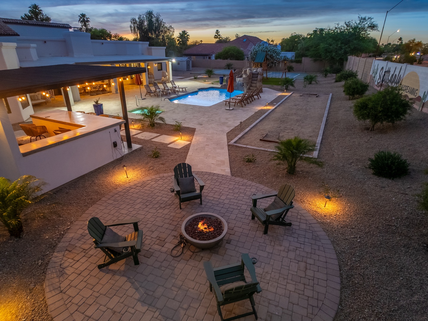 A stunning aerial view showcasing the property's expansive backyard oasis at sunset—featuring a sparkling pool, cozy fire pit seating, covered patio, and beautifully landscaped desert grounds perfect for evening gatherings.