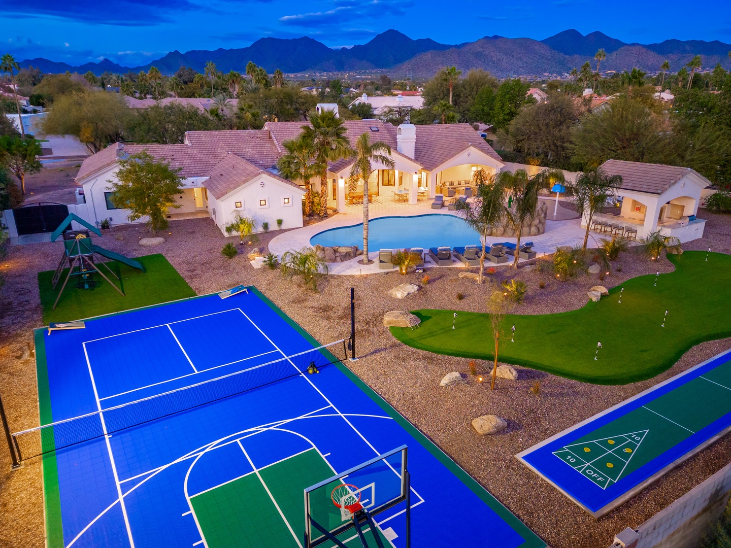 Aerial view of a luxury desert vacation property featuring a private pool, tennis court, miniature golf course, and majestic mountain backdrop at twilight.
