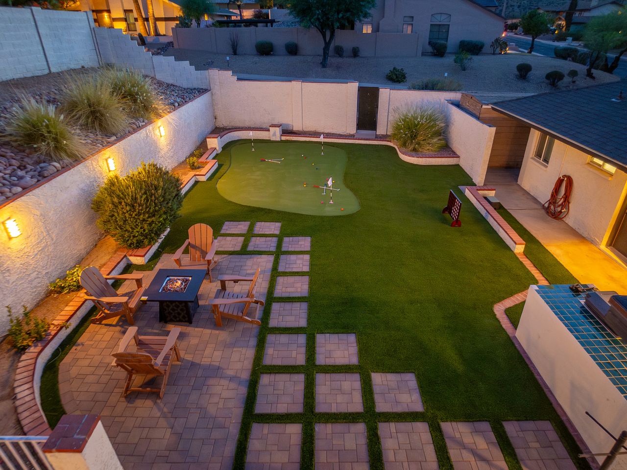 An elevated view of the private backyard at dusk, showcasing a custom putting green, stone patio with fire pit seating, and surrounding neighborhood ambiance illuminated by landscape lighting.