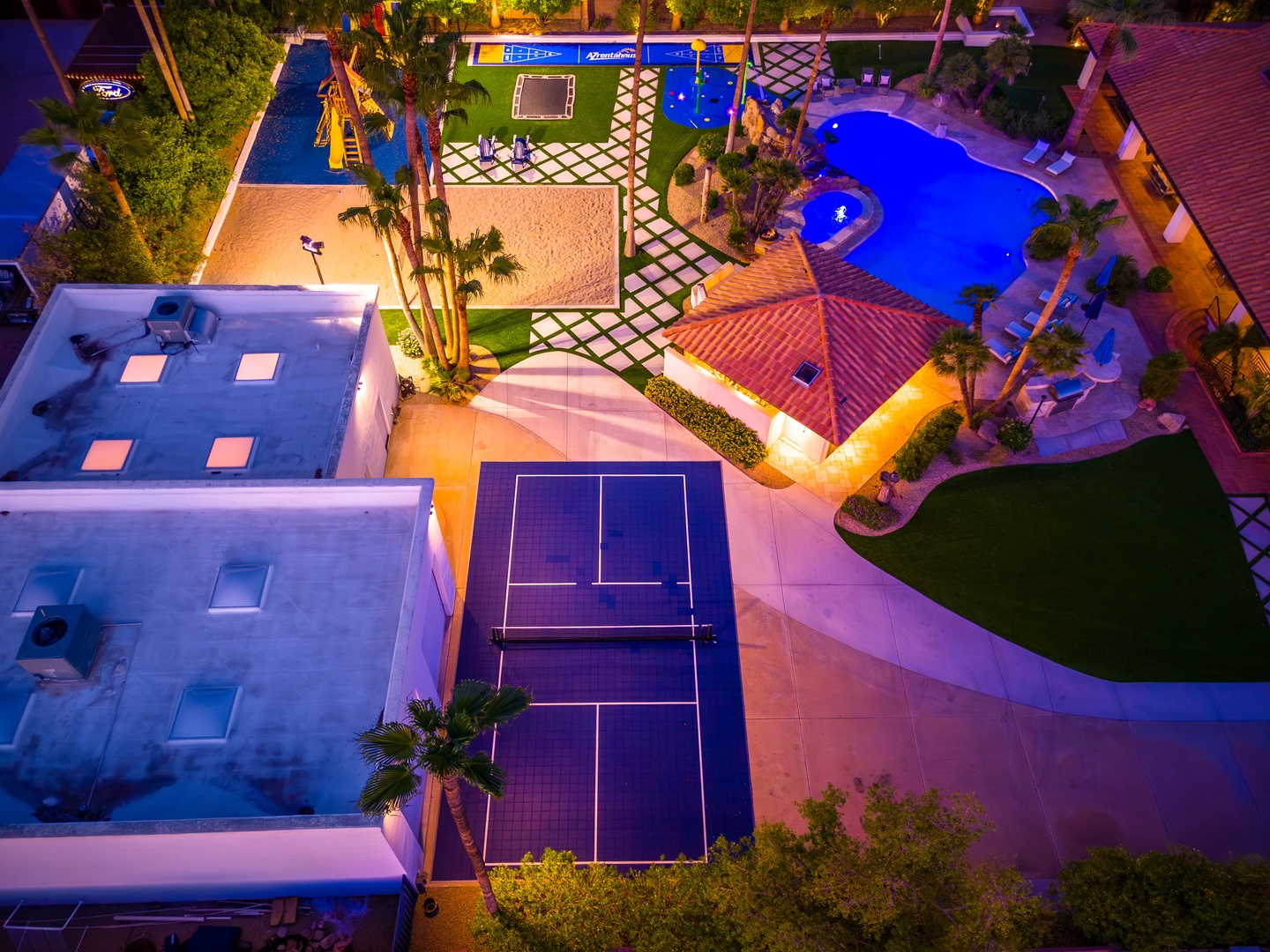 Aerial view reveals a stunning resort-style property featuring multiple swimming pools, a tennis court, lush tropical landscaping, and diverse recreational amenities all illuminated under vibrant evening lighting.