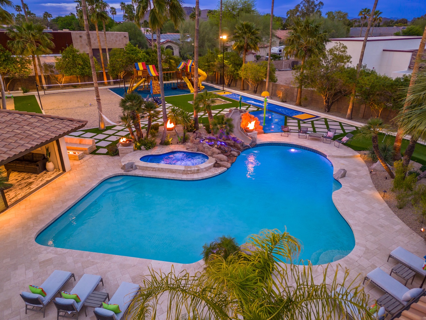 An evening aerial view showcases the property's expansive backyard resort area with multi-level swimming pools, hot tub, water slides, playground equipment, and tropical palm landscaping lit by ambient lighting.