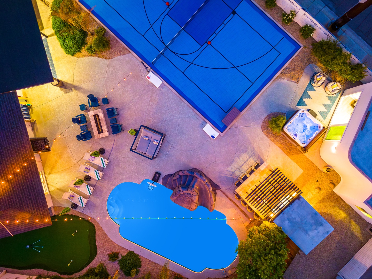 Aerial view of the property showcasing multiple pools, a sports court, hot tub, dining areas, and landscaped outdoor spaces lit for evening ambiance.